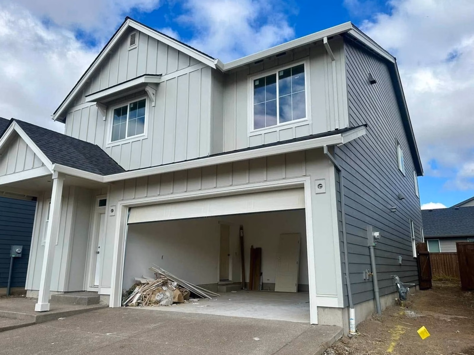 Two-story house with light siding, open garage filled with construction debris, and a partly cloudy sky above.