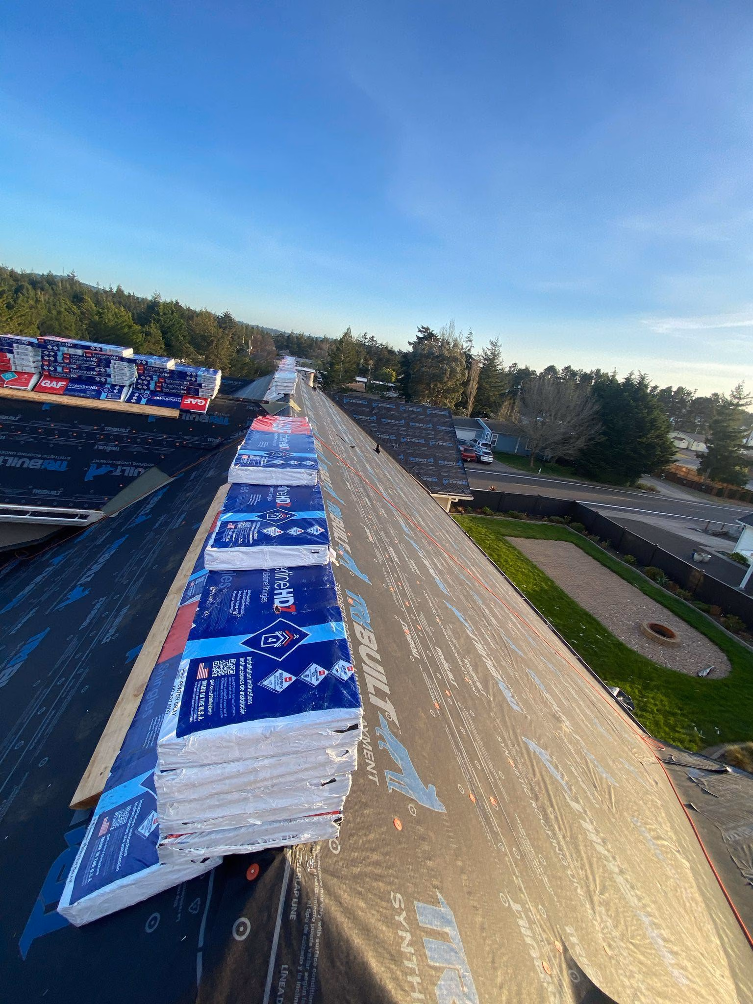Stacks of roofing shingles on a roof under construction with a suburban street and trees in background.
