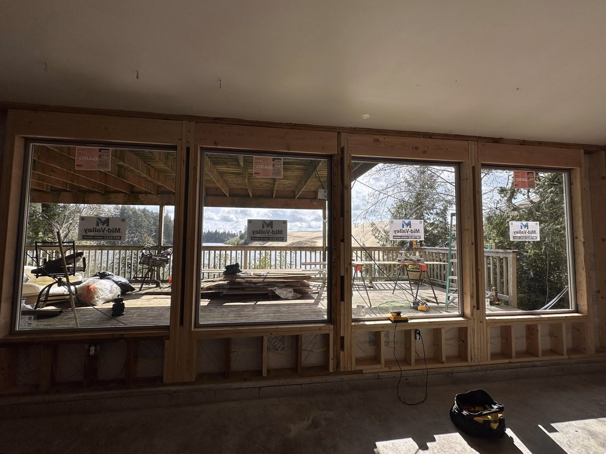 Four large glass windows with wooden frames showing a deck and landscape outside from an unfinished room.