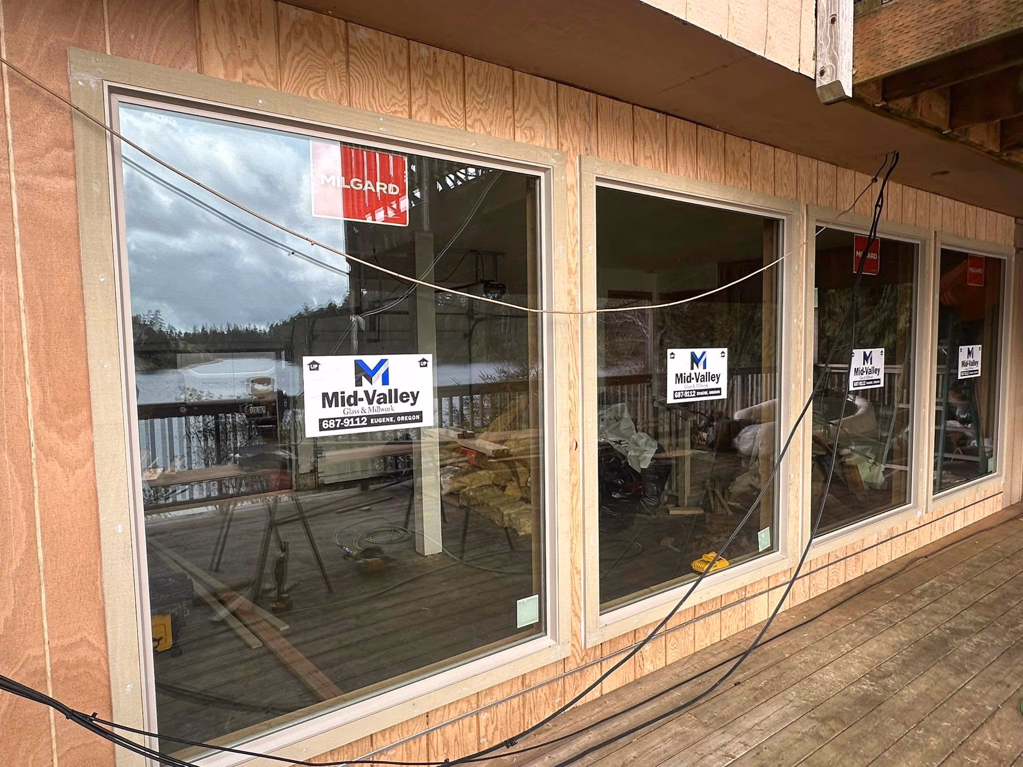 Four large newly installed glass windows on wooden wall overlooking a lake and deck with cables hanging.