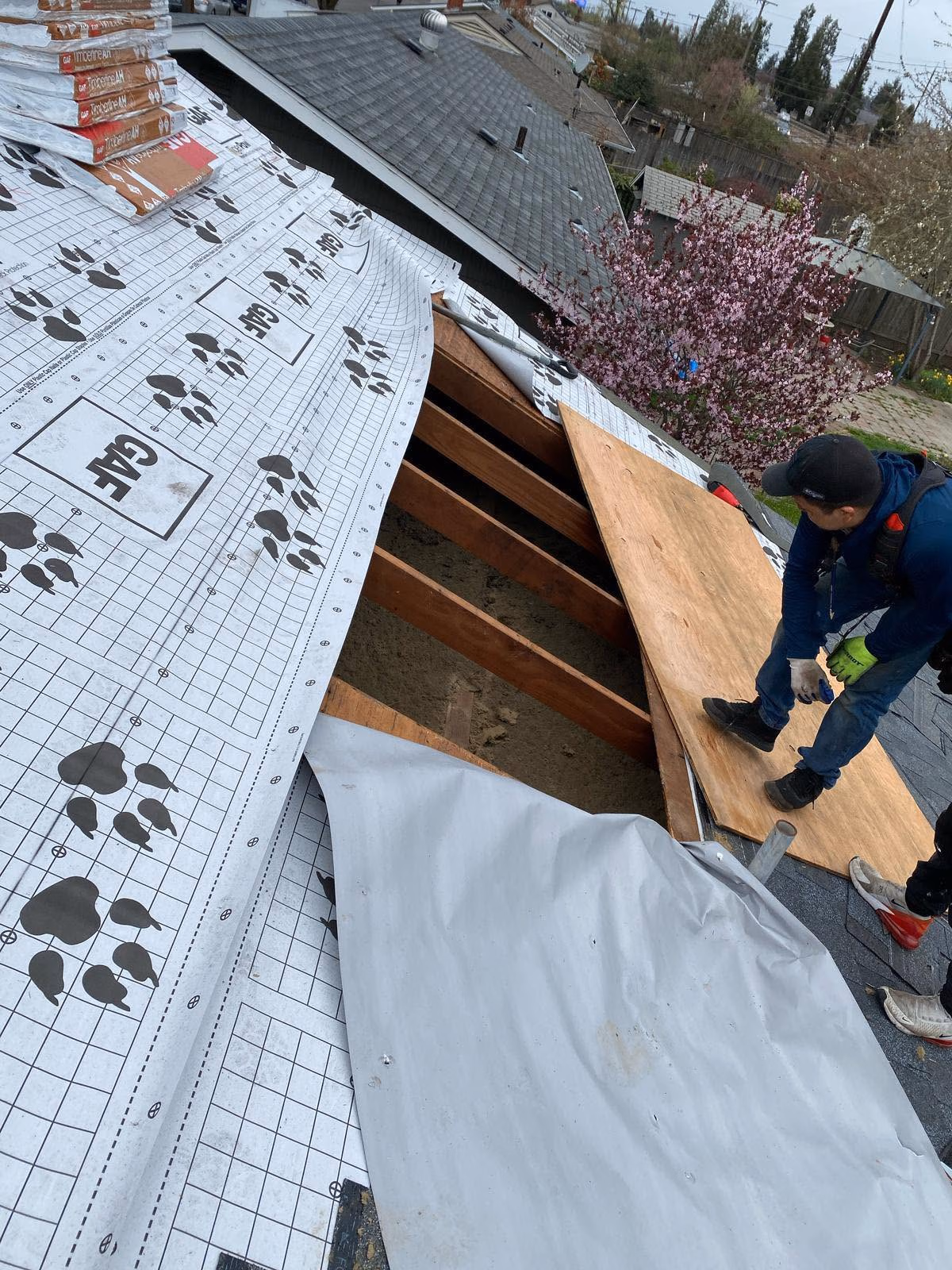 Man in gloves and cap working on roof repair with roofing underlayment and exposed wooden beams.