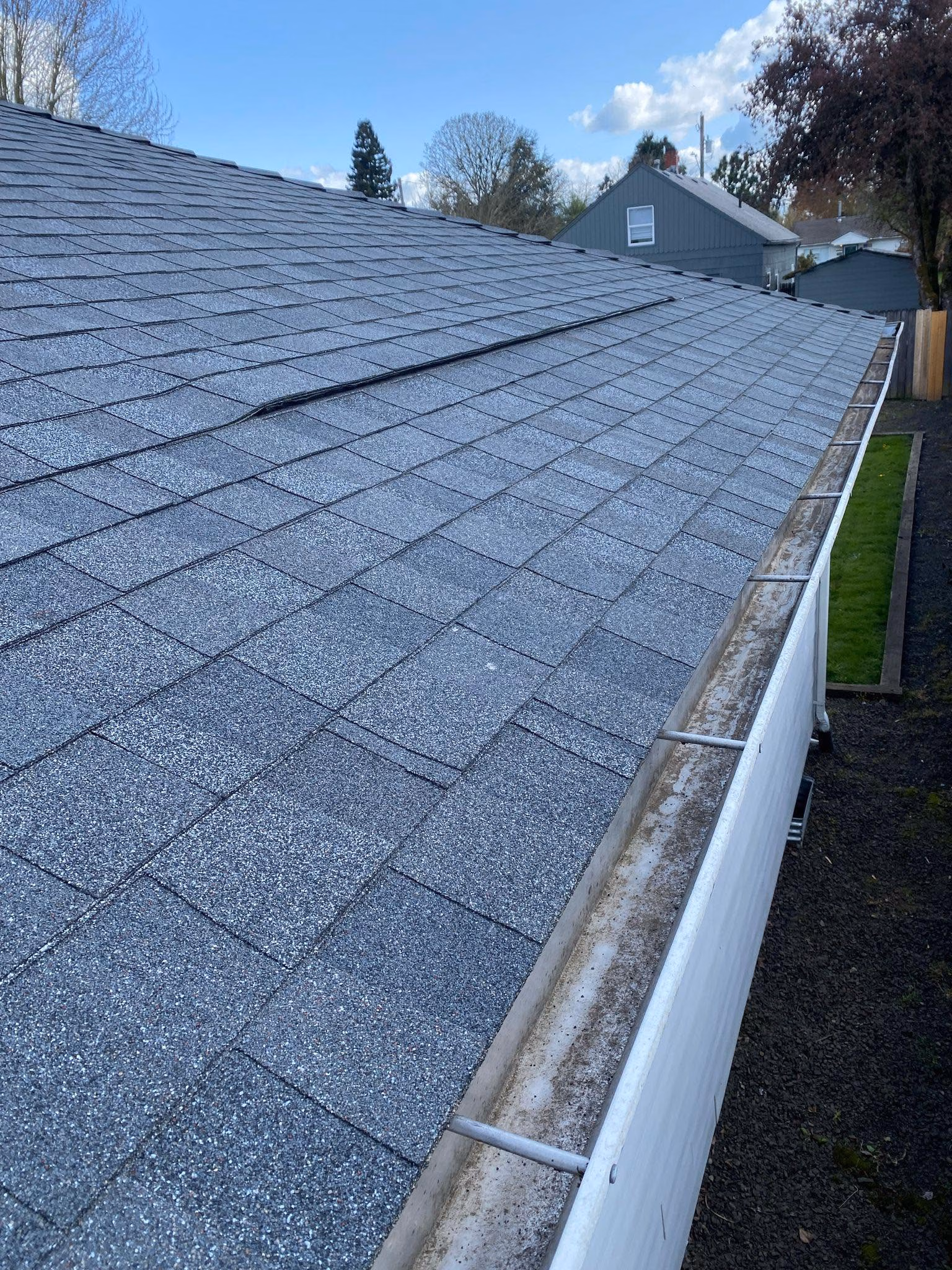 Close-up of a gray shingled roof with a clean white gutter on a sunny day with houses in the background.