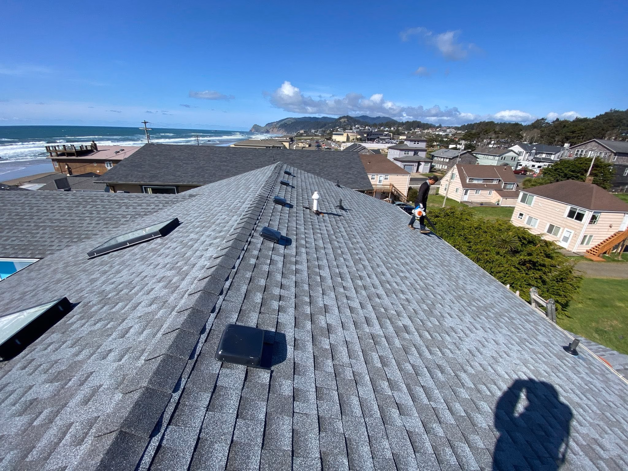 Man walking on a gray shingled roof with coastal houses and ocean in the background under a clear sky.