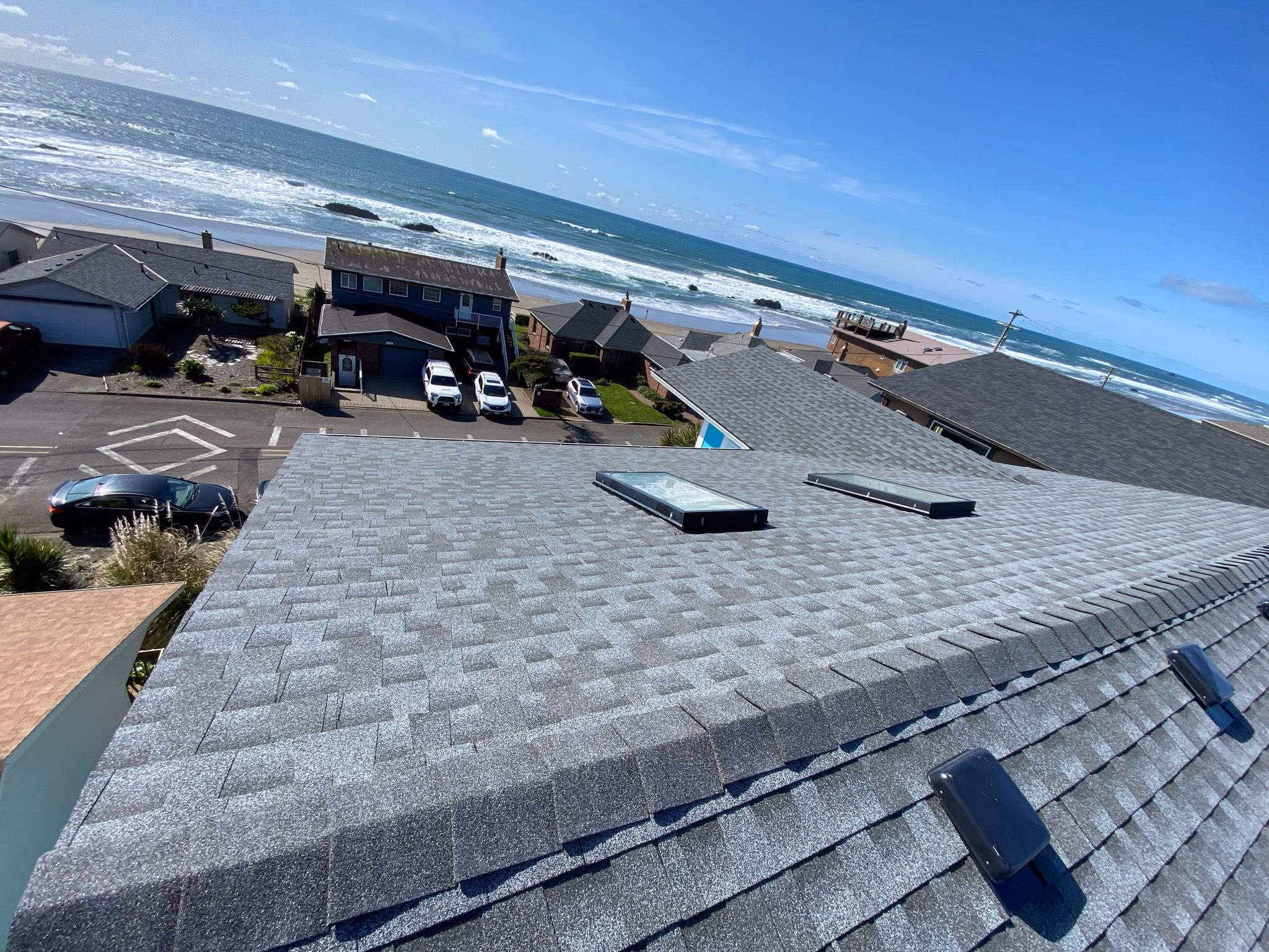 Gray shingled rooftop with two skylights, overlooking parked cars, houses, and an ocean shoreline.