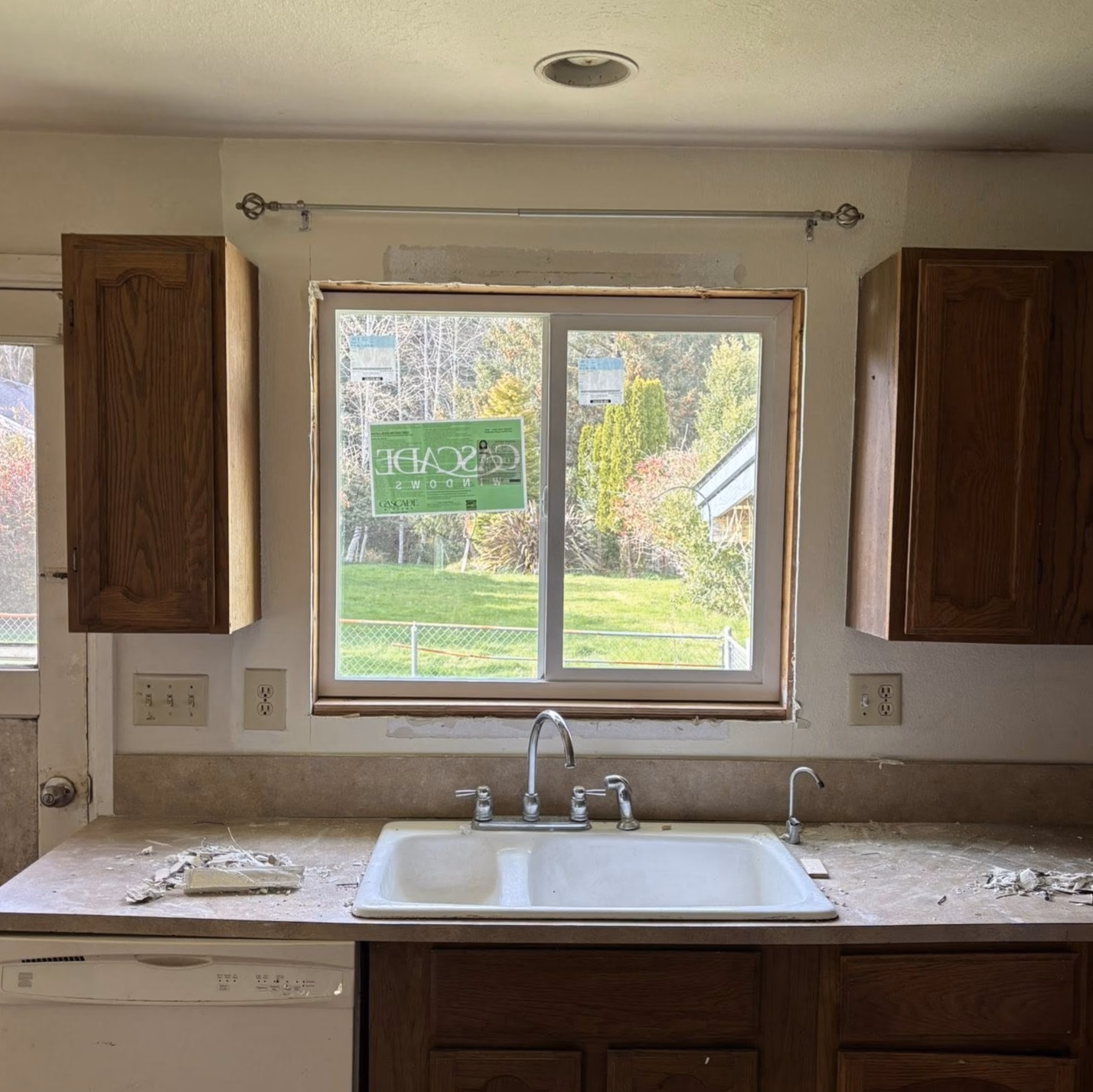 Kitchen sink with dual faucets under a window with Cascade Windows stickers and wooden cabinets on sides.