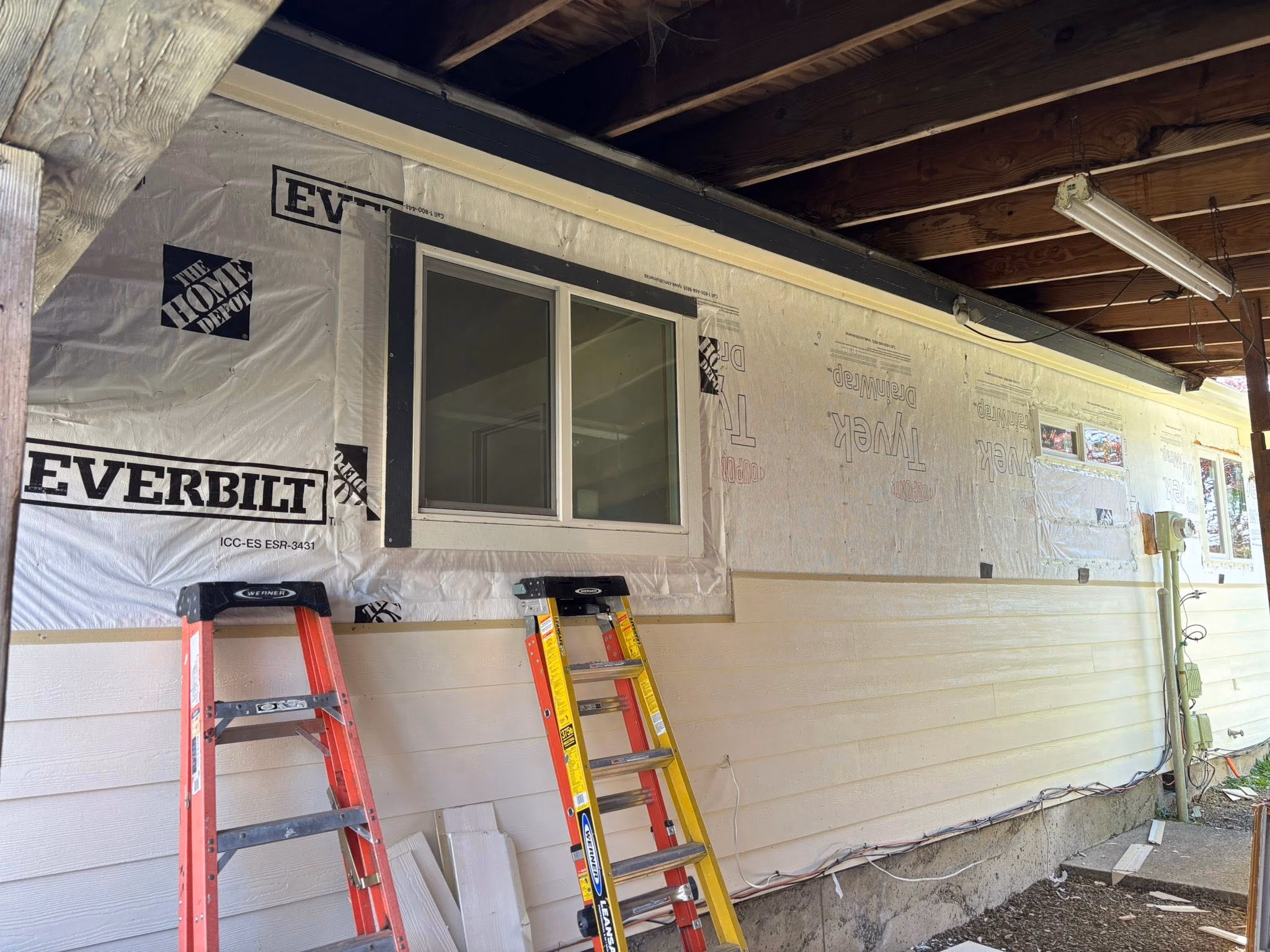 House exterior undergoing siding installation with ladders and covered windows under a wooden ceiling.