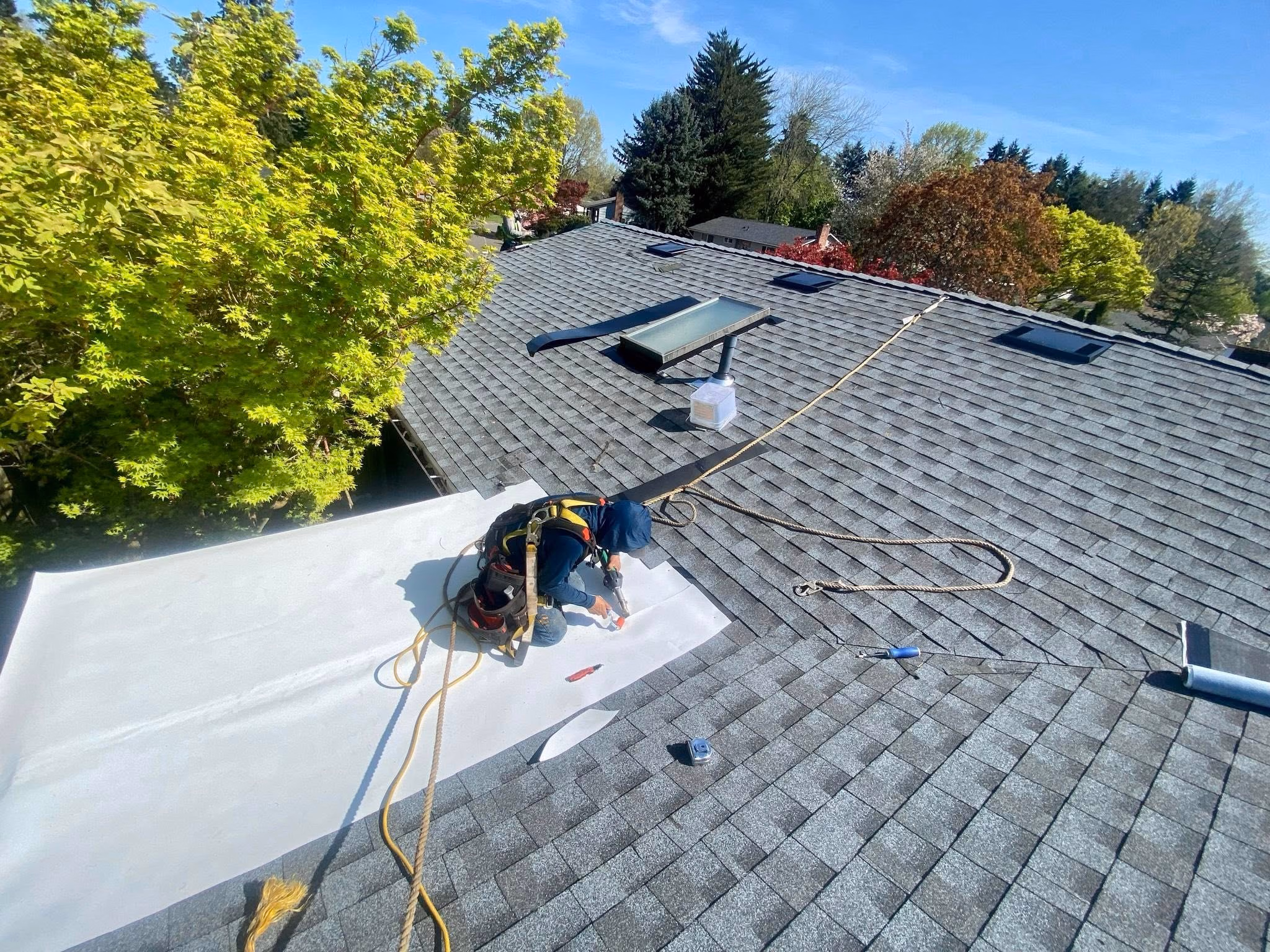 Worker on gray shingled roof installing white roofing membrane with a heat gun and safety harness.
