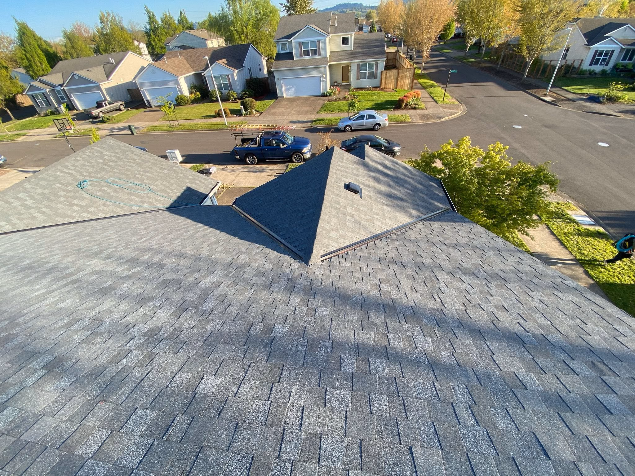 View of gray asphalt shingle roof of a house with neighborhood and cars in the background.