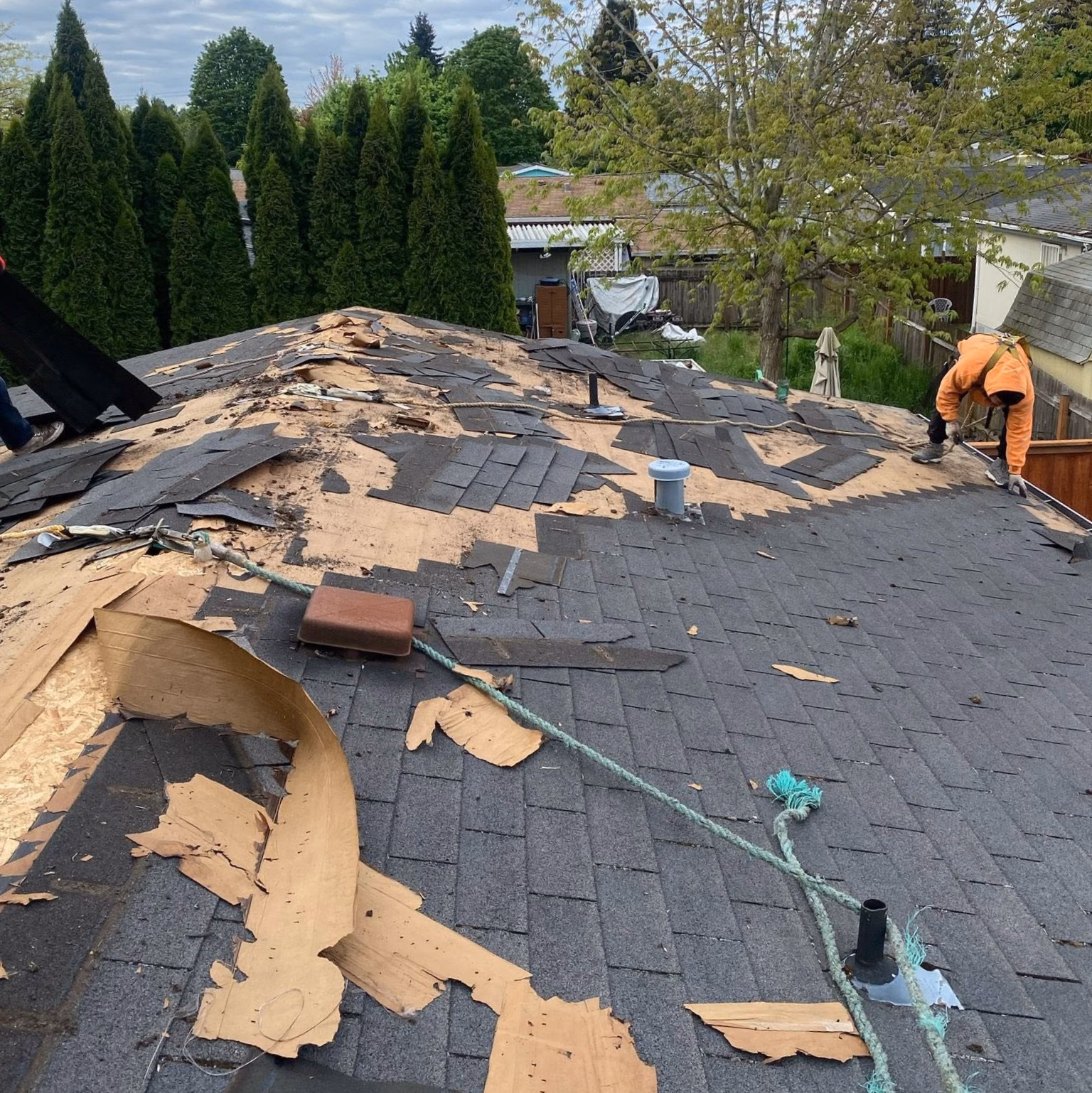 A worker in orange jacket repairing a roof with removed shingles and exposed wood under cloudy sky.