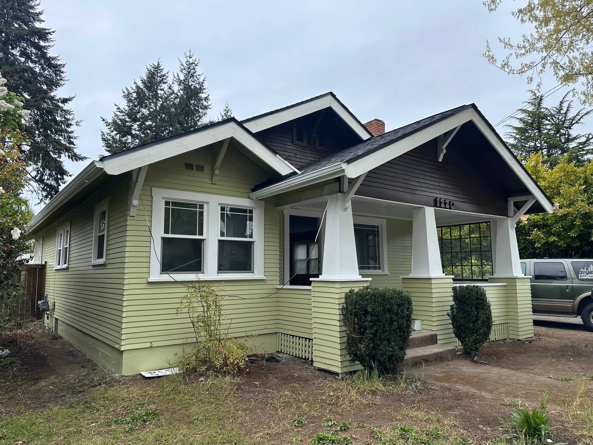 Light green bungalow house with white trim, front porch, and a parked truck nearby under overcast sky.