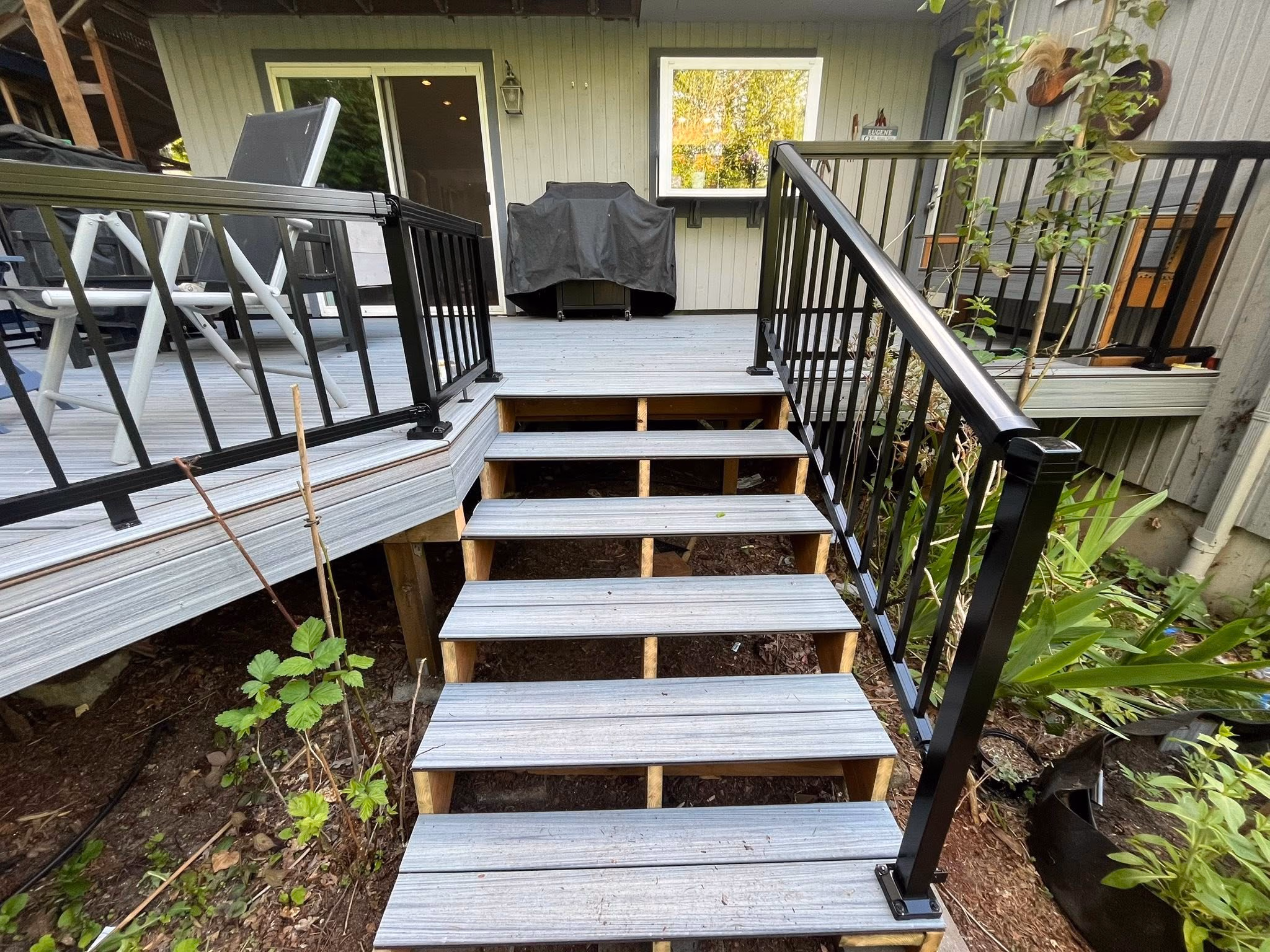 Outdoor wooden stairs with black metal railings leading to a deck with chairs and a covered grill.