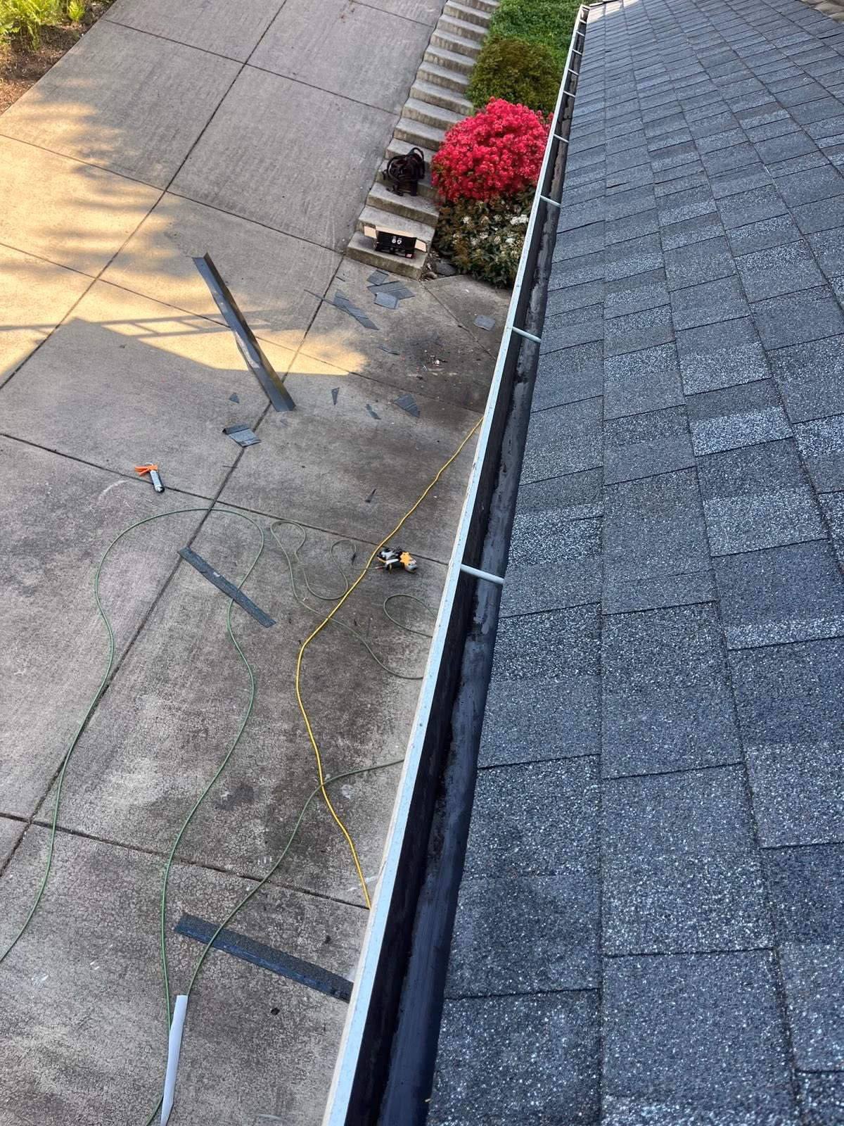 View from roof showing clean gutter, scattered roofing debris, tools, and hoses on driveway below.