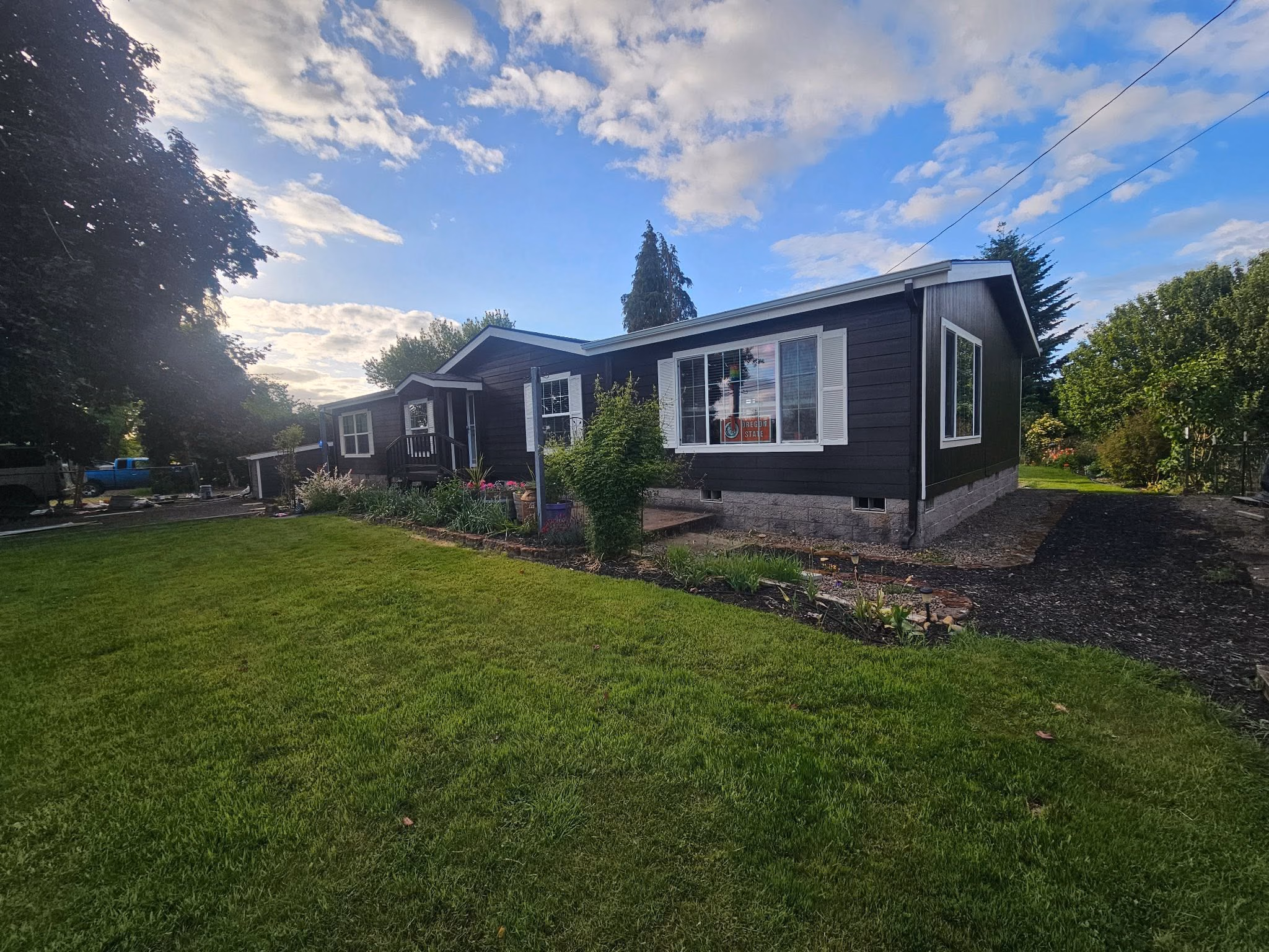 Black single-story house with white window shutters, front lawn, and garden under a partly cloudy sky.