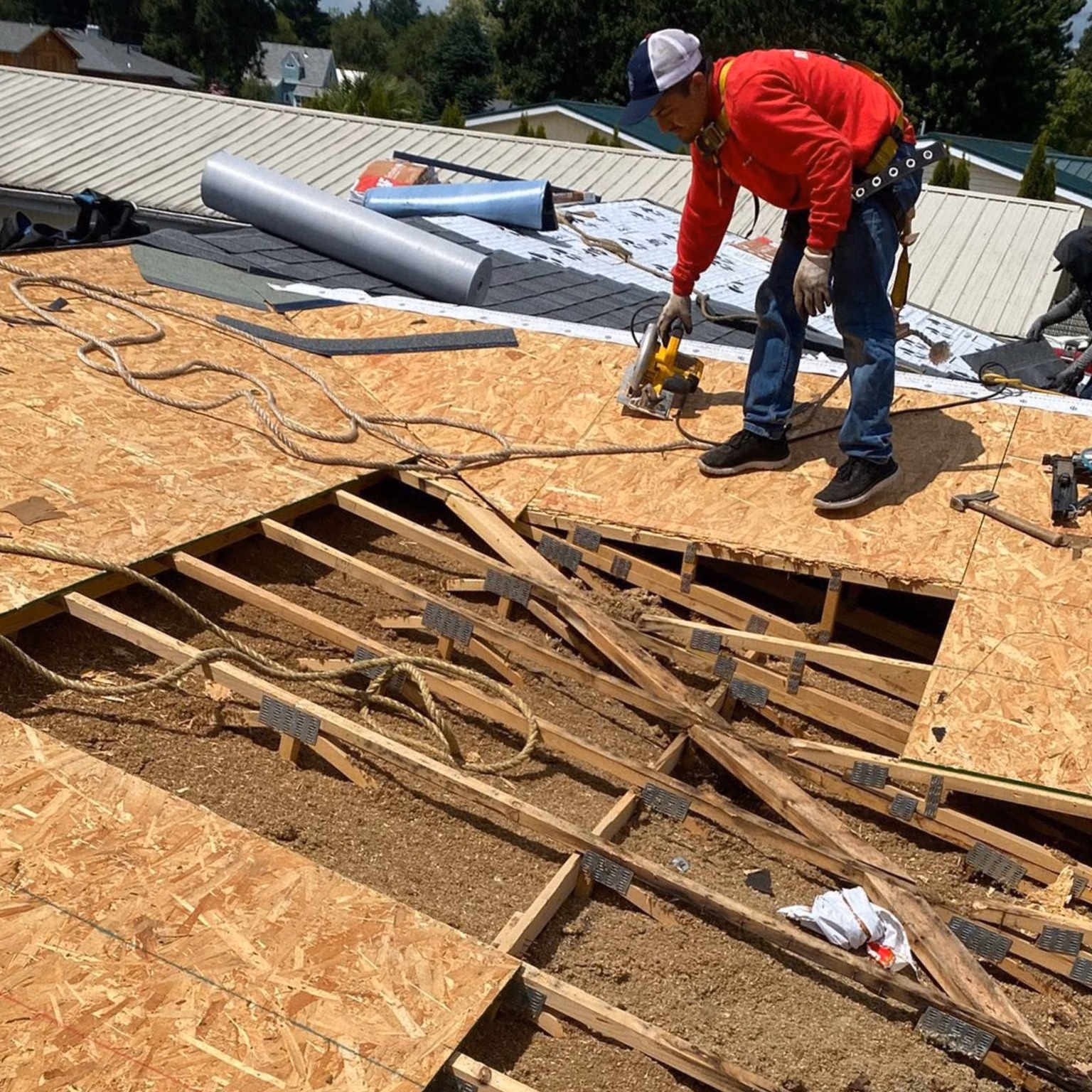 Worker cutting plywood on a roof with exposed wooden beams and roofing materials around.