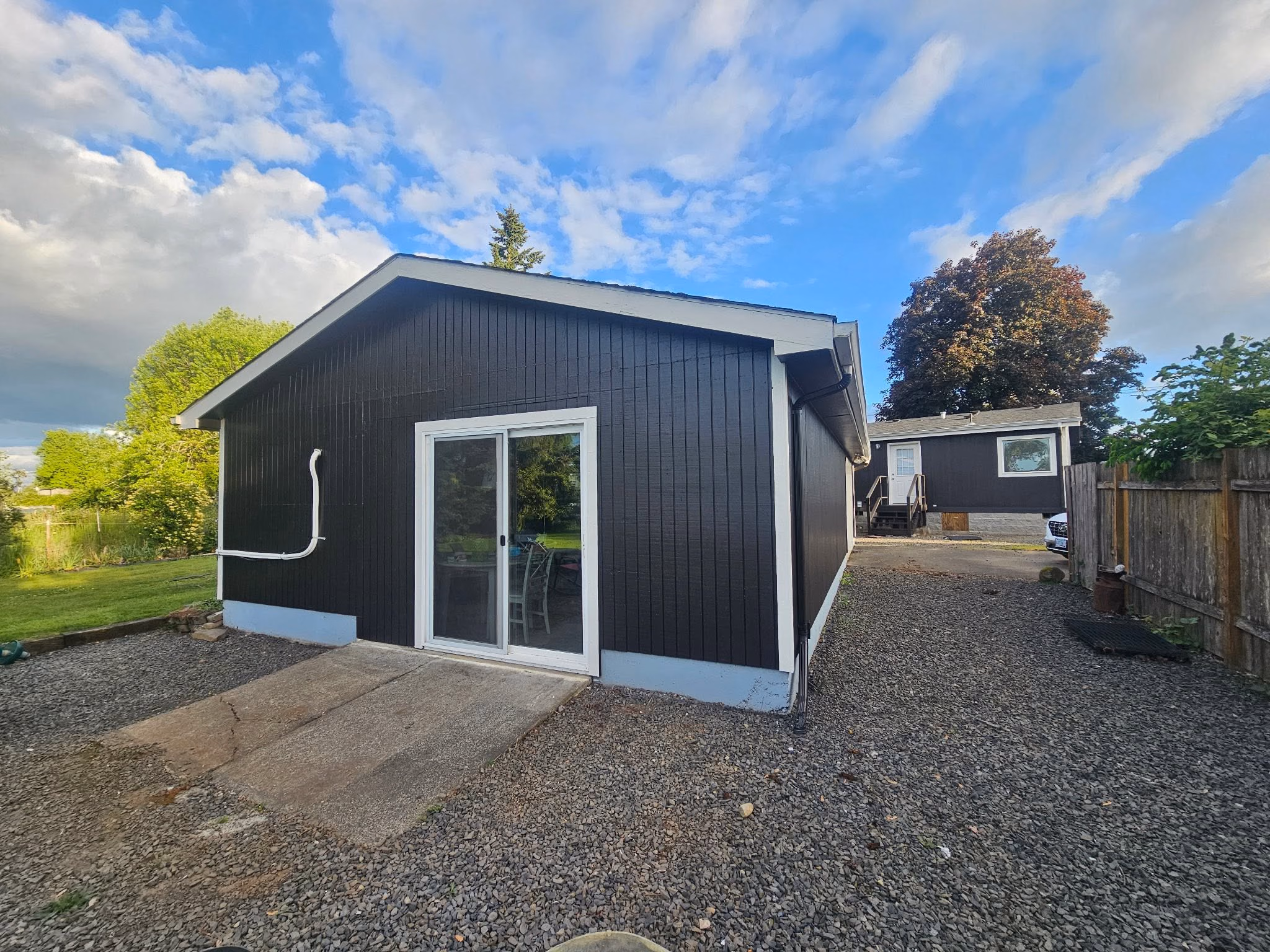 Black house with white trim and sliding glass door beside gravel driveway and green lawn under a partly cloudy sky.