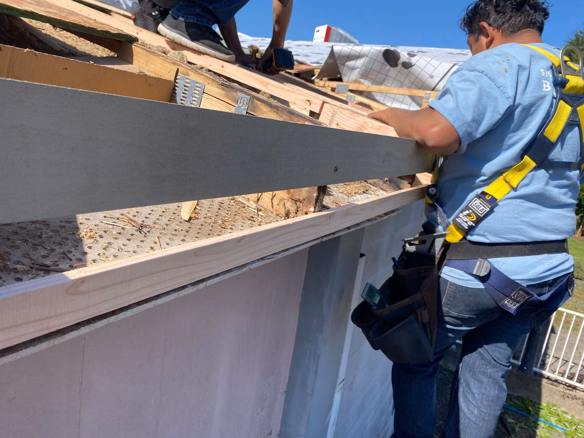 Construction worker in safety harness working on the wooden roof frame under clear blue sky.