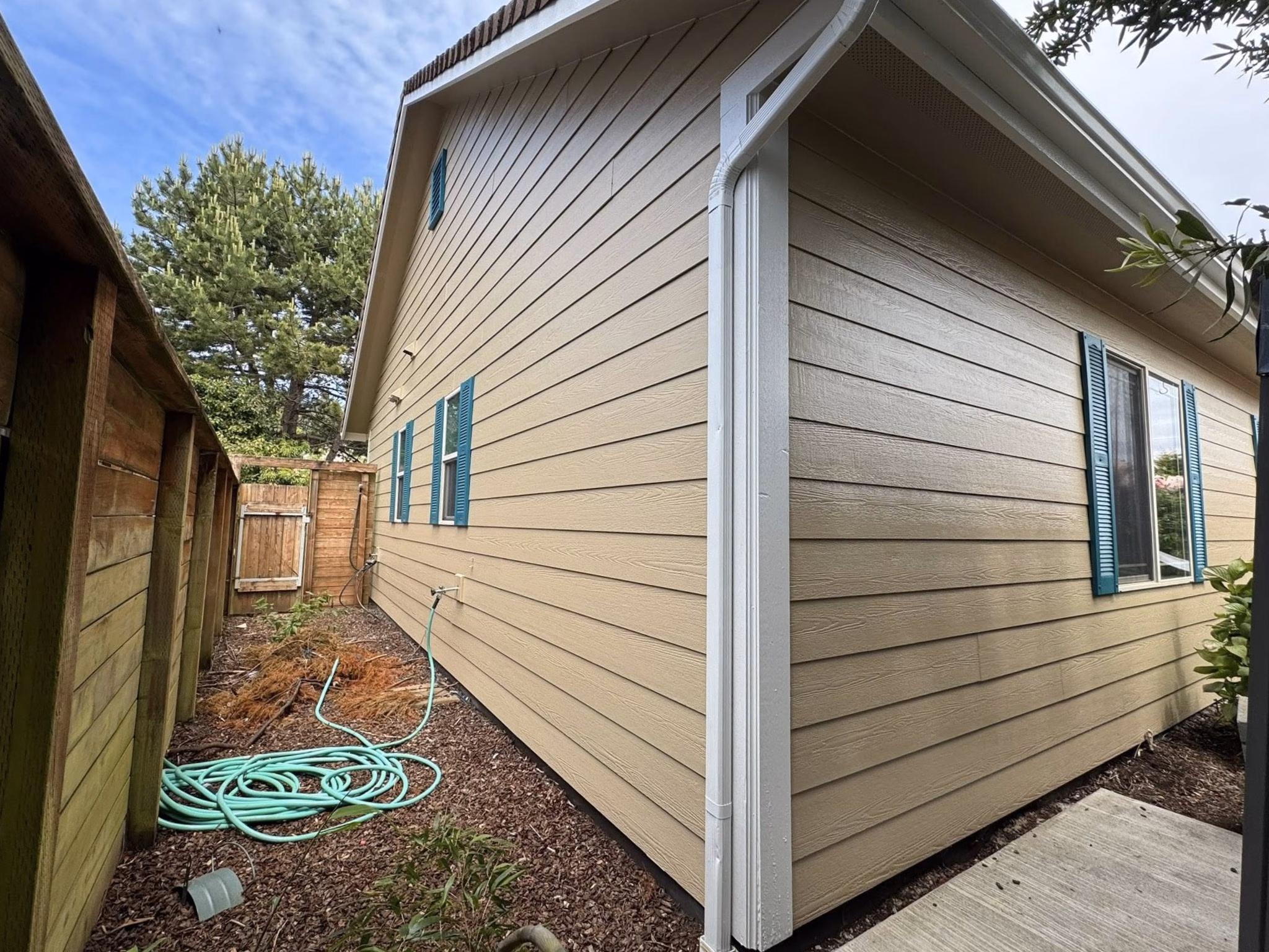 Side view of a beige house with teal window shutters, garden hose, wooden fence, and trees.