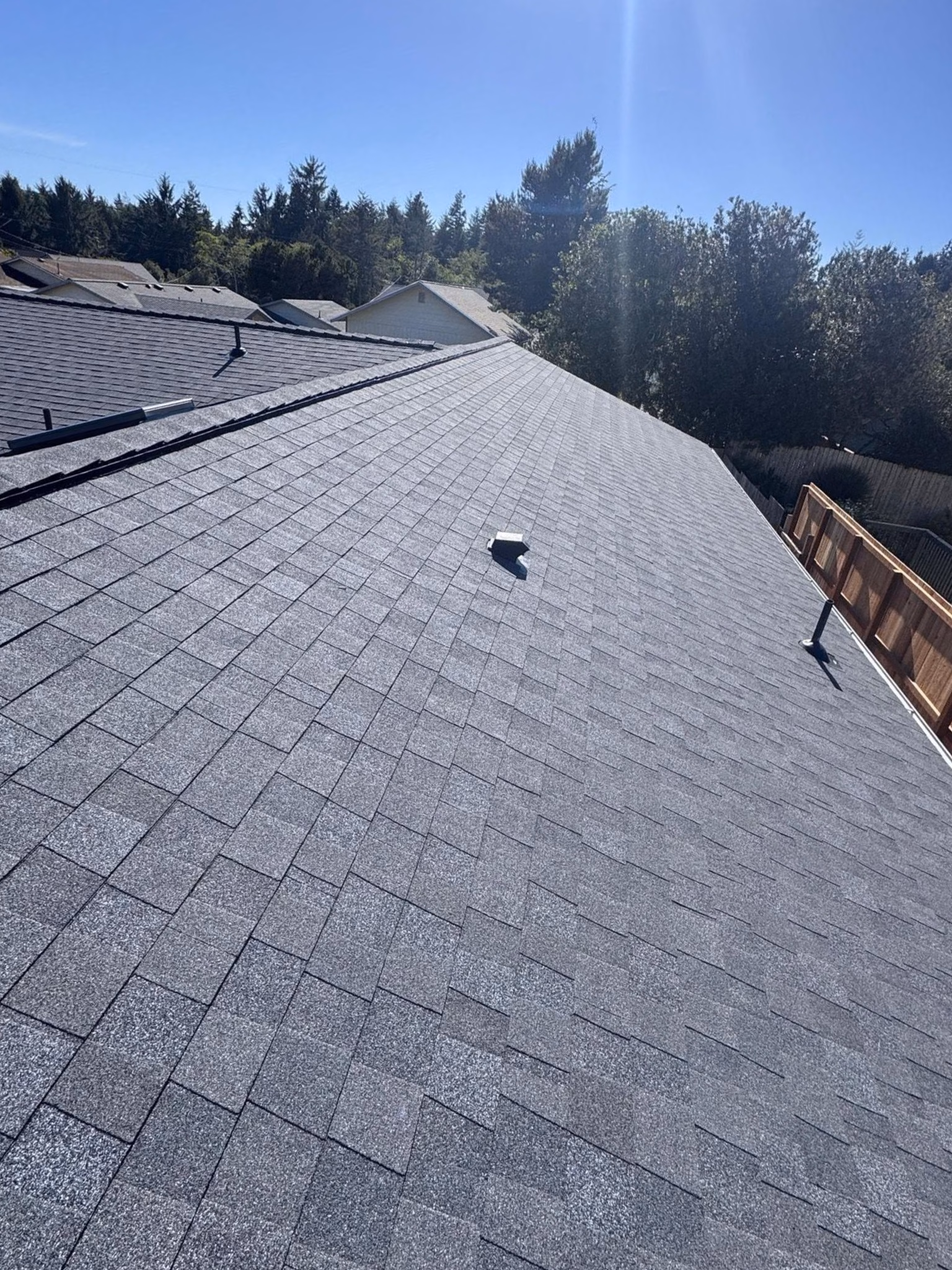 New grey asphalt shingle roof on a house with trees and a clear blue sky in the background.