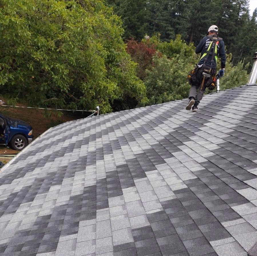 Worker with safety harness walking on gray and black shingled roof surrounded by trees.