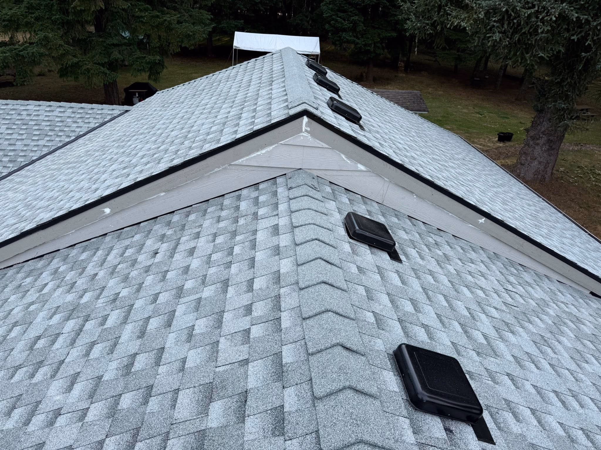 Top view of a gray shingled roof with four black square vents and surrounding trees in the background.