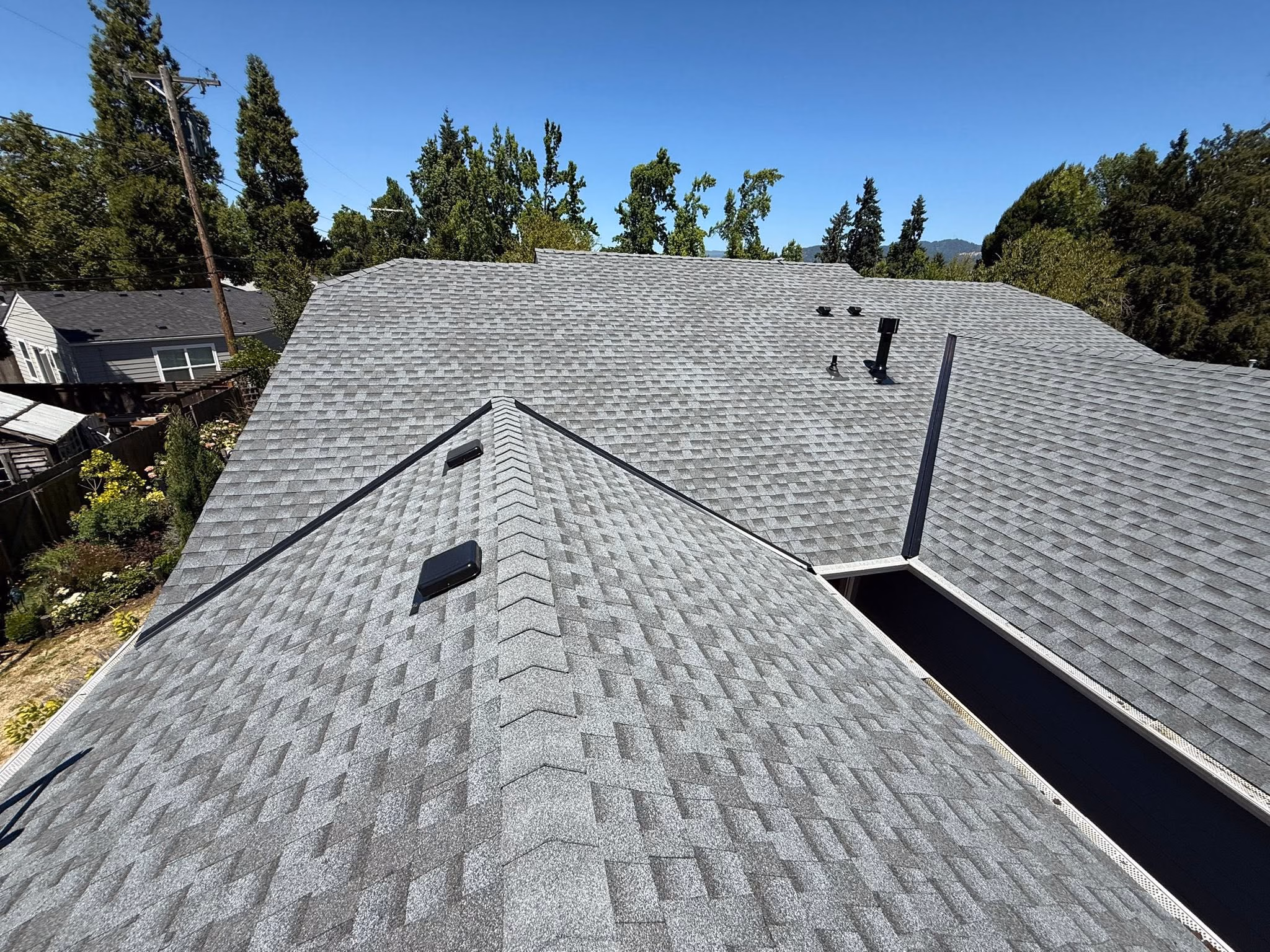 Gray shingled angled rooftops with vents and surrounding green trees under clear blue sky.