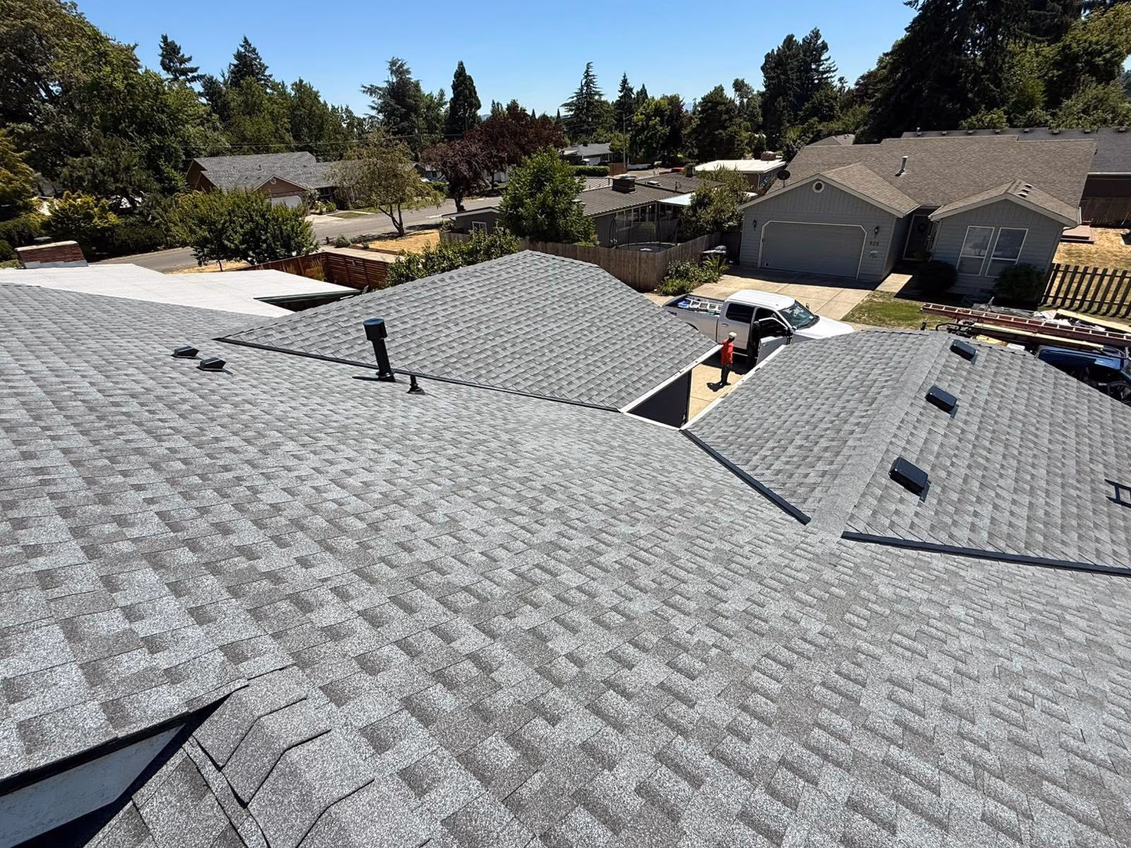 New grey asphalt shingle roofs on suburban houses with trees and trucks visible in the background.