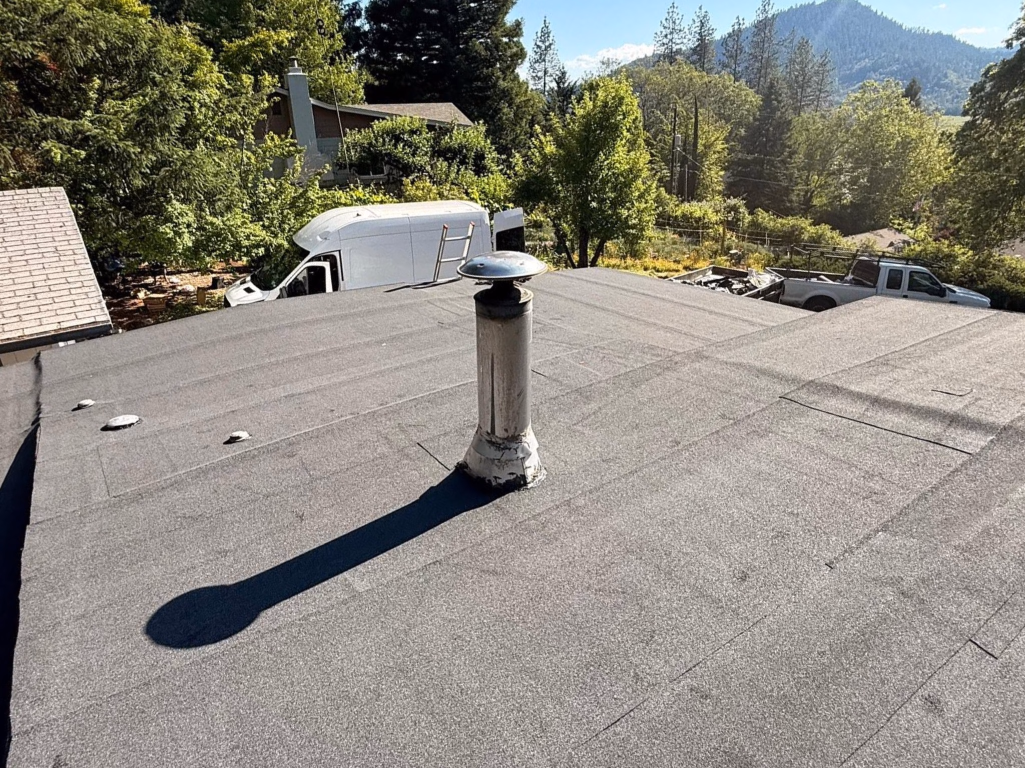 Metal vent pipe casting long shadow on flat gray roof with trees and vehicles in the background.