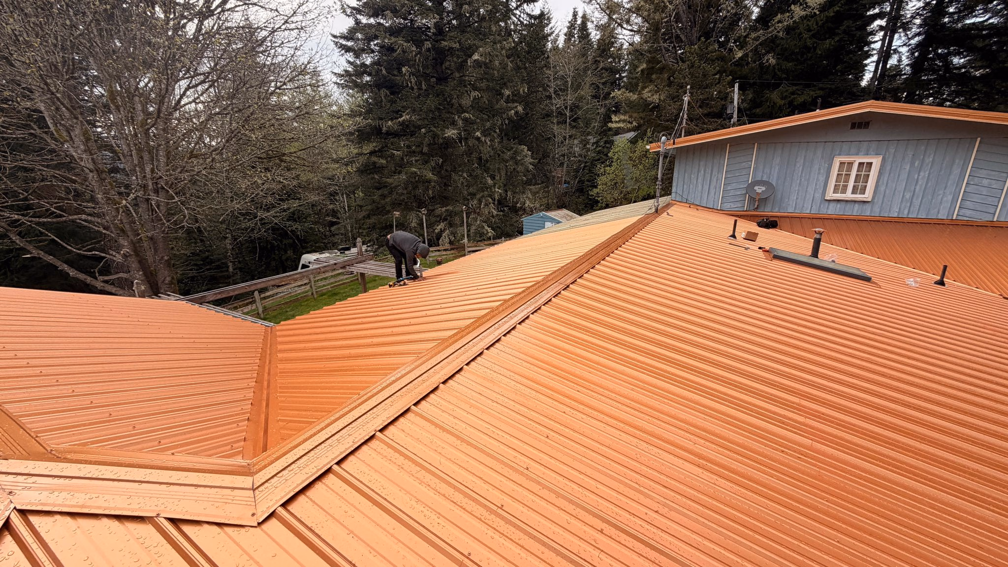 A worker installs or inspects a large orange metal roof on a house surrounded by trees.